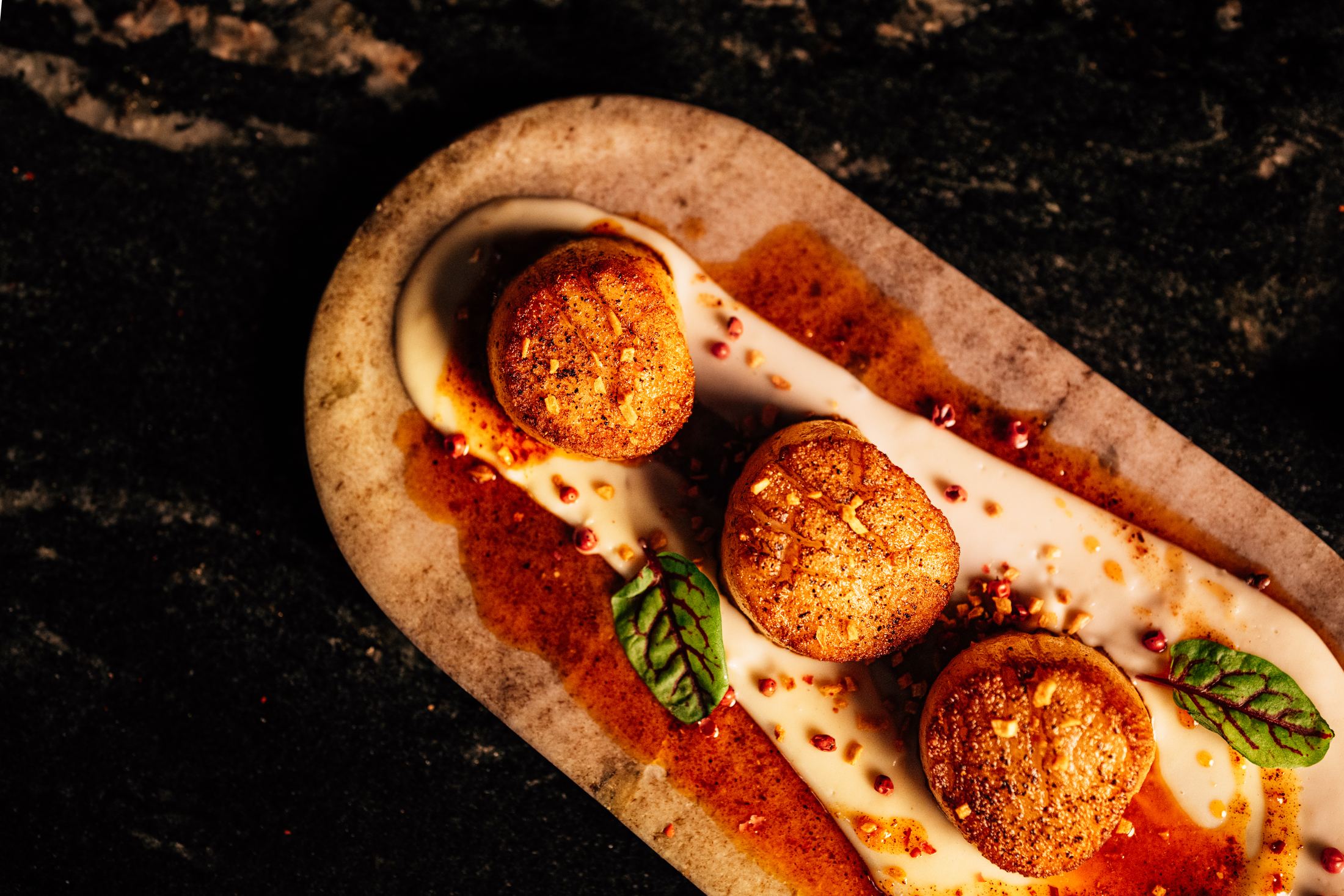 Three seared scallops on a stone board with chili oil and pink peppercorn